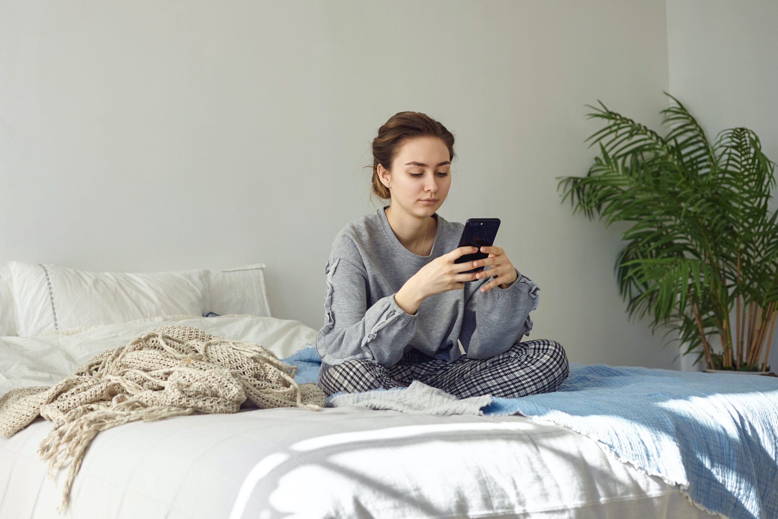 Plan intérieur d'une jolie jeune femme habillée de façon décontractée, assise les jambes croisées sur le lit défait, ayant un regard sérieux tout en lisant un message sur les réseaux sociaux ou en tapant un message texte, à l'aide d'un téléphone portable